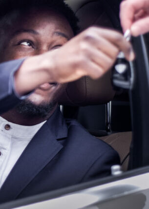 Eine Person sitzt auf dem Fahrersitz eines Autos, und nimmt durch das offene Fenster von einer anderen Person einen Autoschlüssel entgegen