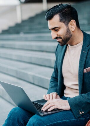 Junger, dunkelhaariger Mann mit gepflegtem Bart und Sakko sitzt auf einer Treppenstufe und schaut auf einen aufgeklappten Laptop, der auf seinen Beinen liegt