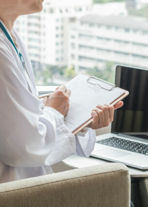 Zwei Personen sitzen sich in Sesseln gegenüber, zwischen ihnen steht ein geöffnetes Laptop und eine der Personen hat ein Klemmbrett in der Hand.