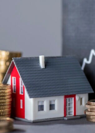 Ein Modellhaus steht zwischen Stapeln von Münzen vor einer Tafel mit einer steigenden Kurve.