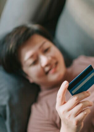 Eine Person liegt auf der Couch. Sie hält in der einen Hand eine Kreditkarte und in der anderen ein Smartphone.