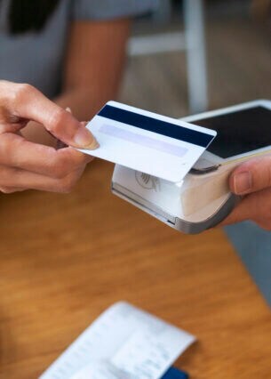 Eine Person hält eine Bankkarte an ein elektrisches Lesegerät, das eine andere Person hält. Auf einem Tisch darunter liegt ein Kassenbon.
