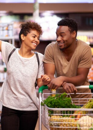 Zwei Personen zwischen Supermarktregalen. Sie schieben einen mit Gemüse gefüllten Einkaufswagen.