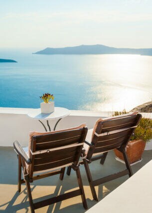 Zwei Holzstühle stehen auf einer sonnigen Terrasse mit Blick übers Meer.