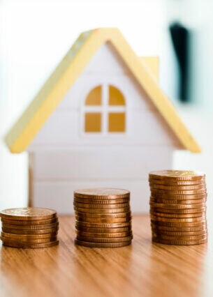 Ein weißes Miniaturhaus. Davor drei unterschiedlich hohe Stapel Geldmünzen.