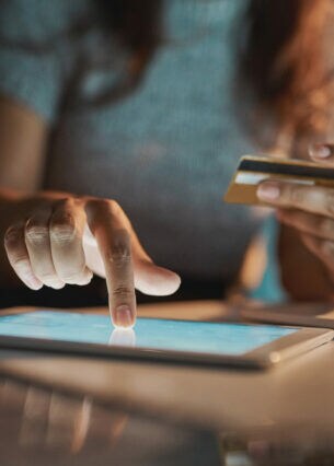 Eine Person sitzt mit Bezahlkarte in der einen Hand am Tablet und tippt mit der anderen Hand auf den Bildschirm