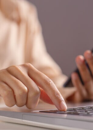 Eine Frau hält ein Handy in der linken Hand und drückt mit der rechten Hand eine Taste auf ihrem Laptop