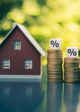 Ein Modellhaus neben vier Stapeln mit Münzen. Auf den Münzstapeln, die von links nach rechts immer kleiner werden, ist jeweils ein Würfel mit Prozentzeichen platziert