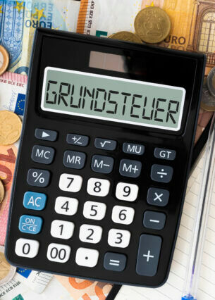 Ein Taschenrechner liegt auf Euroscheinen und Euromünzen, auf dem Display steht das Wort Grundsteuer.