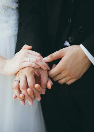 Nahaufnahme von Händen mit Eheringen. Sie gehören einer Frau im Brautkleid sowie einem Mann im Anzug.