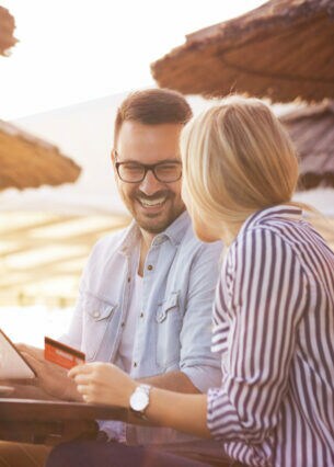 Junges Paar sitzt lachend unter Strandschirmen mit Tablet und Kreditkarte.