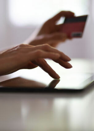 Nahaufnahme einer Hand, die auf einem Tablet tippt. Im Hintergrund ist eine zweite Hand zu erkenne, die eine Bankkarte in der Hand hält.