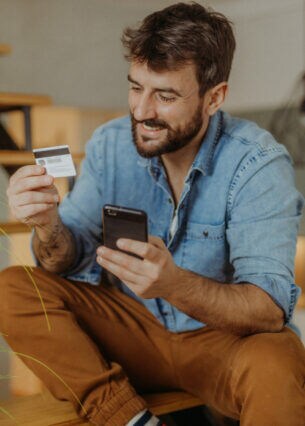 Ein Mann sitzt lächelnd auf den Stufen einer Treppe. In der einen Hand hält er eine Kreditkarte, auf die er schaut. In der anderen Hand hält er ein Smartphone