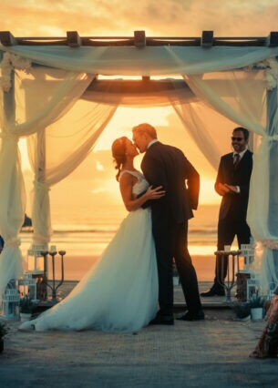 Ein Brautpaar steht bei Sonnenuntergang unter einem Baldachin im Freien und küsst sich. Mehrere Gäste sitzen auf Stühlen und klatschen