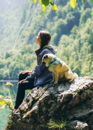 Eine Person und ein kleiner Hund sitzten zusammen auf einem Felsen vor einem See, der von Wald umsäumt ist