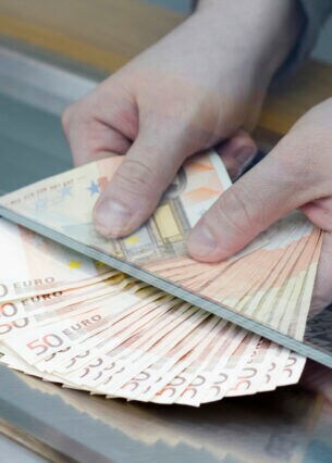 Eine Person reicht einige aufgefächerte 50-Euro-Banknoten unter einer Glaswand hindurch.
