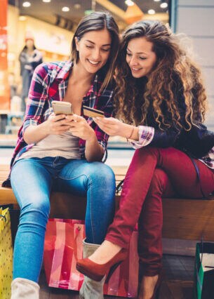Zwei Personen sitzen in einer Shoppingmall auf einer Bank und schauen gemeinsam auf ein Smartphone und eine Kreditkarte.