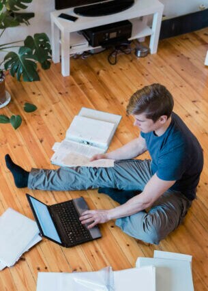 Eine Person sitzt auf dem Fußboden mit Aktenordnern und einem Laptop und wird von oben fotografiert.