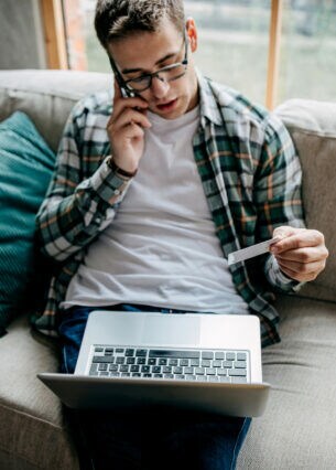 Eine Person aus der Aufsicht, die telefonierend auf einem Sofa sitzt, eine Bankkarte in der Hand hält und ein Laptop auf dem Schoß liegen hat.