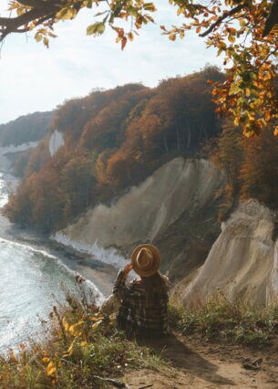Eine Person mit großem Hut sitzt an einer Klippe mit Blick Richtung Meer.