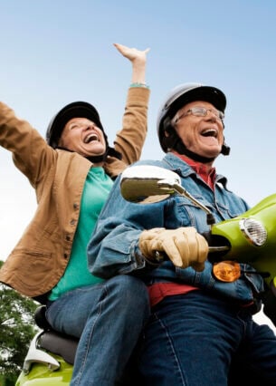 Ein älterer Mann und eine ältere Frau sitzen lachend auf einem grünen Moped. Die Frau streckt beide Arme in die Höhe.