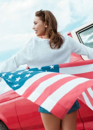Eine weibliche Person in Rückansicht vor einem roten Auto, die eine amerikanische Flagge hinter sich wehen lässt.