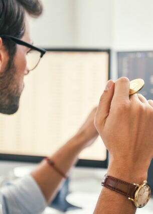Ein Mann schaut auf den Kursverlauf auf seinem Computermonitor und hält eine Münze in der Hand