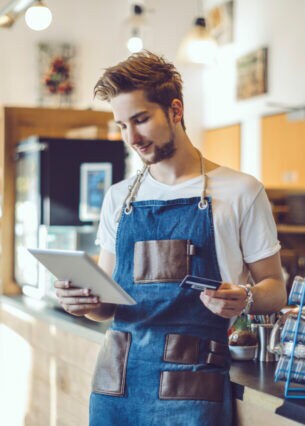 Ein junger Barista steht in einem Café, er hat eine Schürze an und hält in der einen Hand ein Tablet und in der anderen Hand eine Kreditkarte