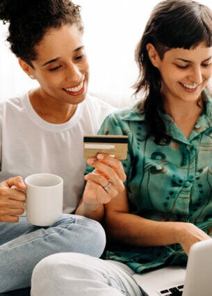 Zwei auf dem Sofa sitzende Personen halten eine Tasse und eine Kreditkarte in der Hand. Eine Person hat einen Laptop auf dem Schoß.