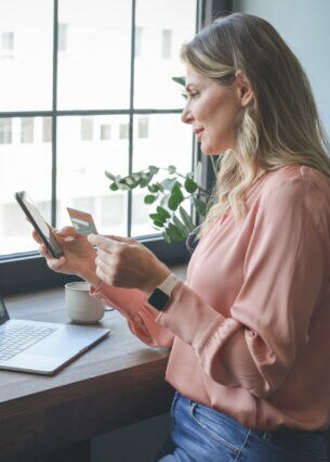 Eine Frau steht mit Handy und Kreditkarte in der Hand vor einem Tisch, auf dem ein Laptop steht