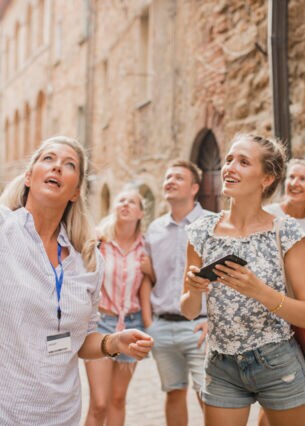 Eine Gruppe von Menschen steht in der Straße einer Altstadt. Sie hören einer Reiseleiterin zu.