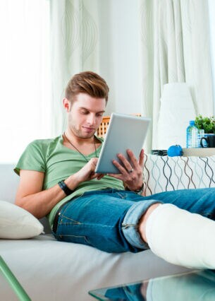 Eine Person mit gebrochenem Bein sitzt auf einem Sofa und schaut auf sein Tablet, neben ihr stehen Krücken.