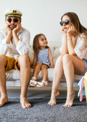 Eine Familie sitzt auf einem Sofa und stützt die Köpfe auf ihre Hände, daneben stehen gepackte Koffer.