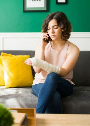 Eine Person sitzt mit einbandagiertem Arm auf einem Sofa und telefoniert.
