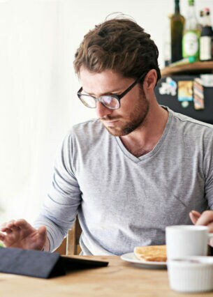 Eine Person sitzt am Frühstückstisch und tippt auf ihr Tablet.