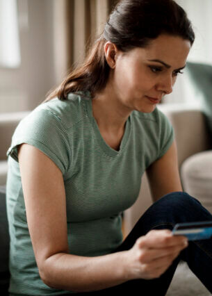 Eine Person sitzt auf dem Fußboden vor einem Sofa, hält in der einen Hand ein Smartphone und in der anderen Hand eine Bankkarte.