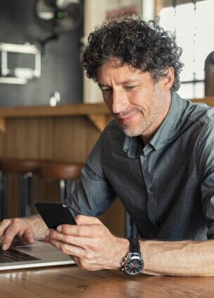 Eine Person blickt auf sein Smartphone und sitzt vor einem geöffneten Laptop.
