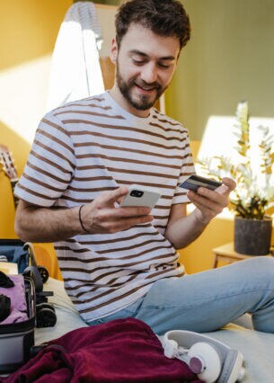 Eine Person sitzt neben einem aufgeklappten Koffer auf dem Bett und hält eine Bankkarte sowie ein Smartphone in den Händen.