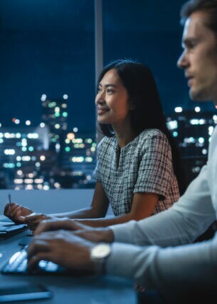 Zwei Personen sitzen in einem Büro in einer Großstadt am Schreibtisch, auf den vier Bildschirmen sind Börsencharts, Indikatoren und weitere Daten zu sehen.