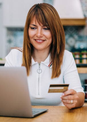 Eine Frau sitzt an einem Tisch vor einem aufgeklappten Laptop und hält eine Kreditkarte in der Hand.