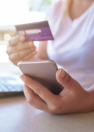 Eine Person sitzt am Tisch vor einem aufgeklappten Notebook. In Händen hält sie Smartphone und Kreditkarte.