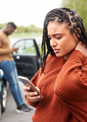 Zwei Autos stehen hintereinander auf einer Straße. Ein Mann lehnt am hinteren Auto, im Vordergrund sieht eine junge Frau besorgt auf ihr Smartphone.