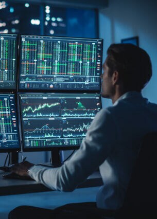 Zwei Personen sitzen jeweils an einem Schreibtisch vor mehreren Bildschirmen mit Kursverläufen und Zahlentabellen.