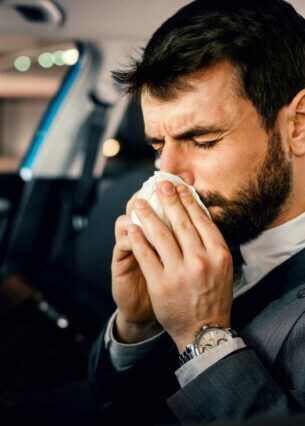 Ein dunkelhaariger Mann im Anzug sitzt angeschnallt am Steuer eines parkenden Autos und putzt seine Nase.