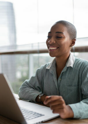 Eine lächelnde Person sitzt an einem Tisch vor einem aufgeklappten Laptop.
