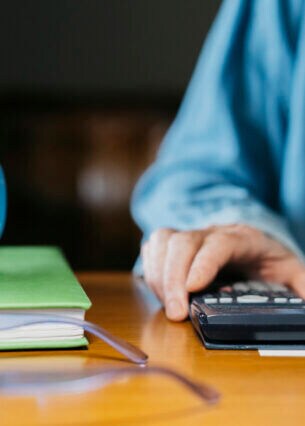 Eine Person an einem Tisch hält ihre Hand auf einen Taschenrechner, der neben einem Notizbuch und einer Tasse liegt.