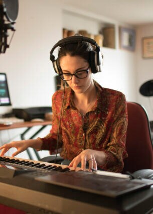 Eine Person sitzt in ihrer Wohnung an einem Keyboard, trägt Kopfhörer und ist von technischem Musikequipment umgeben.