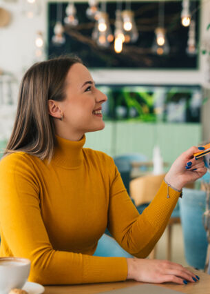 Eine Frau im gelben Rollkragenpullover an einem Tisch im Café mit einer Kaffeetasse hält eine Bezahlkarte an das Kartenlesegerät der Bedienung.