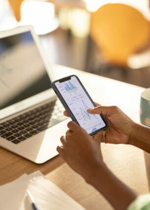 Eine Person sitzt mit Kaffee und Laptop an einem Tisch und studiert einen Aktienkurs auf dem Smartphone.