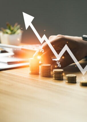 Eine Person an einem Tisch hält in einer Hand ein Smartphone, mit der anderen Hand stapelt sie Geld. Die Geldstapel sind mit einem nach oben steigenden Kursverlauf illustriert.